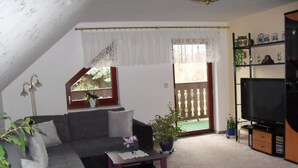 Sala de estar con un sofá modular gris, una mesa de centro, un mueble para televisión y una estantería. Se ve una ventana con vistas al exterior, una puerta y una planta en el alféizar.