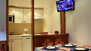 Zona pranzo con tavolo apparecchiato per quattro persone, televisore a parete e cucina sullo sfondo.