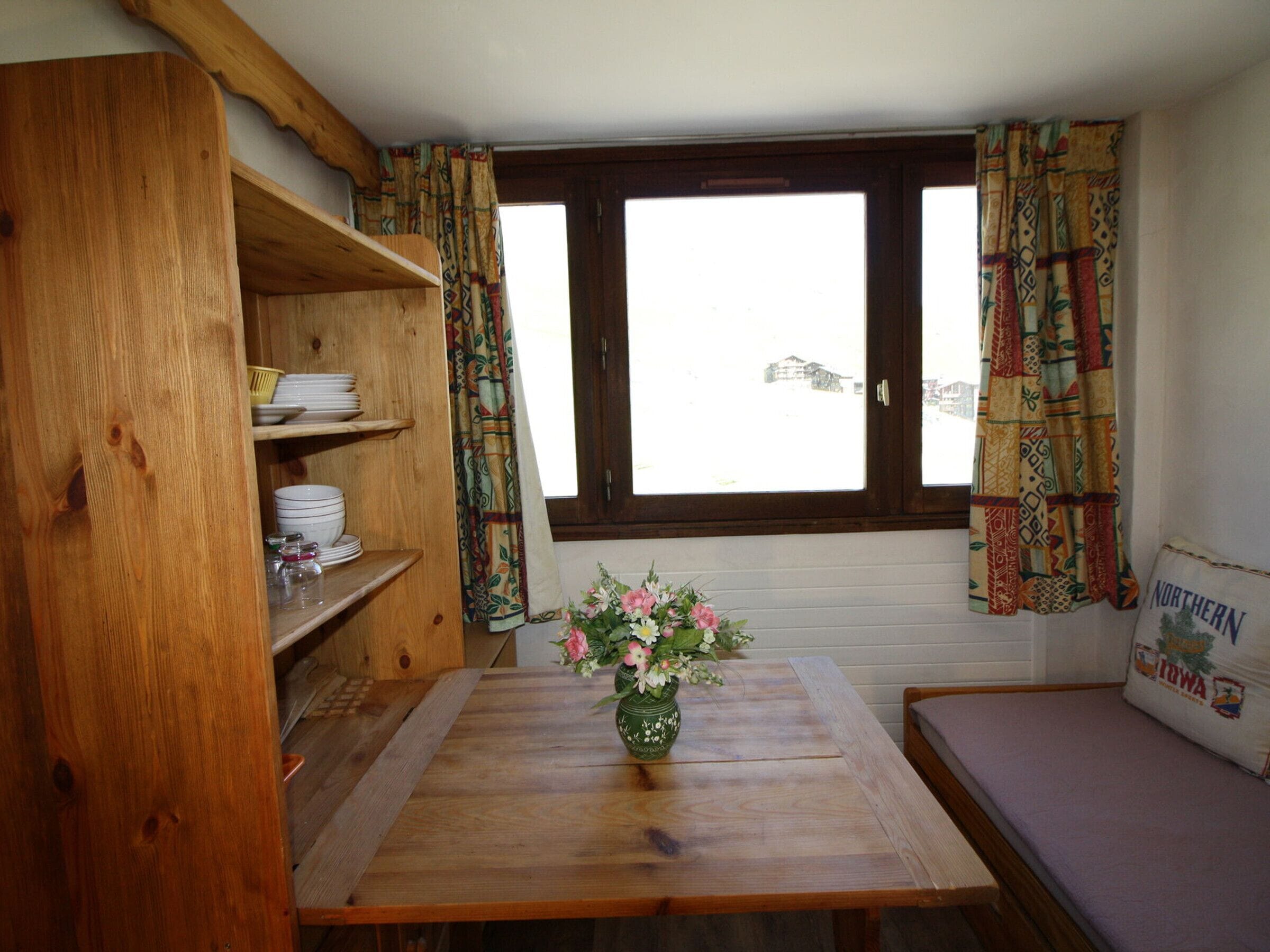 Une table à manger en bois ornée d’un vase de fleurs, une étagère en bois avec de la vaisselle et une fenêtre avec des rideaux.