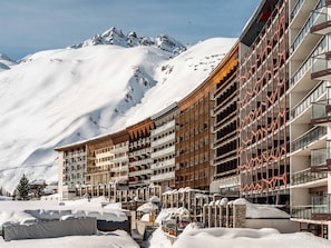 A hotel with multiple balconies, a ski slope in the background, and snow-covered surroundings.