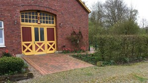 A brick building with a yellow door and a small garden area with flowers and a wooden fence. - Dirkshof 2 West, Germany (Varel)