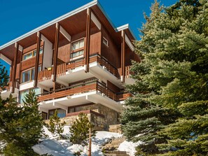 A multi-story building with balconies, stone foundation, and surrounding evergreen trees.