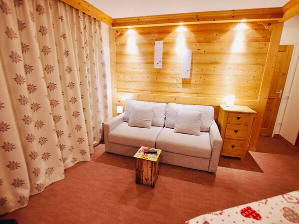 Een knus ingerichte kamer met een houten hoofdbord, een sofa met veel kussens, een nachtkastje en een klein houten bijzettafeltje met een tijdschrift.