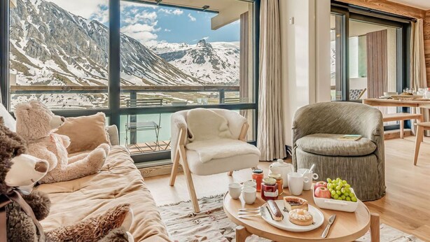 Un accogliente soggiorno con una grande finestra che si affaccia su un paesaggio montano innevato, un divano confortevole, una poltrona e un tavolino con snack e bevande.