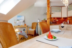 Een eetgedeelte met een tafel voor twee personen, een keuken met houten kasten en uitzicht op het strand door een raam.