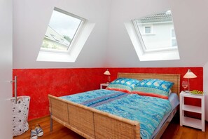 A bedroom with a red wall, a bed with blue patterned bedding, two bedside tables with lamps, and a window.