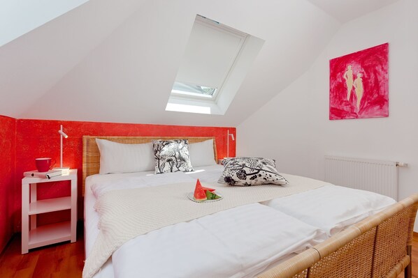 Ein Schlafzimmer mit schrägem Dach, einem Oberlicht, einem Bett mit weißer Bettwäsche, einer roten Akzentwand und einem Gemälde.