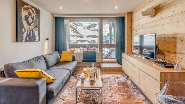 A modern living room with a sofa, coffee table, and a TV. There is a large window with a view of mountains.