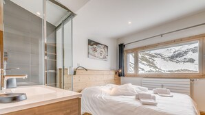 A modern bedroom with a wooden headboard, a large window with a mountain view, a white bed, and a bathroom with a glass shower enclosure.