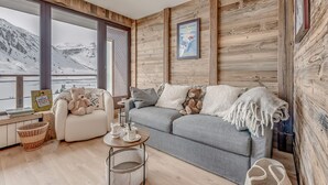 A cozy living room with a grey sofa, a white armchair, and a small table with a teapot and cups. A large window offers a view of snowy mountains. - Premium 3-room apartment, lake view, 6 pers., WIFI, in Tignes (Tignes)