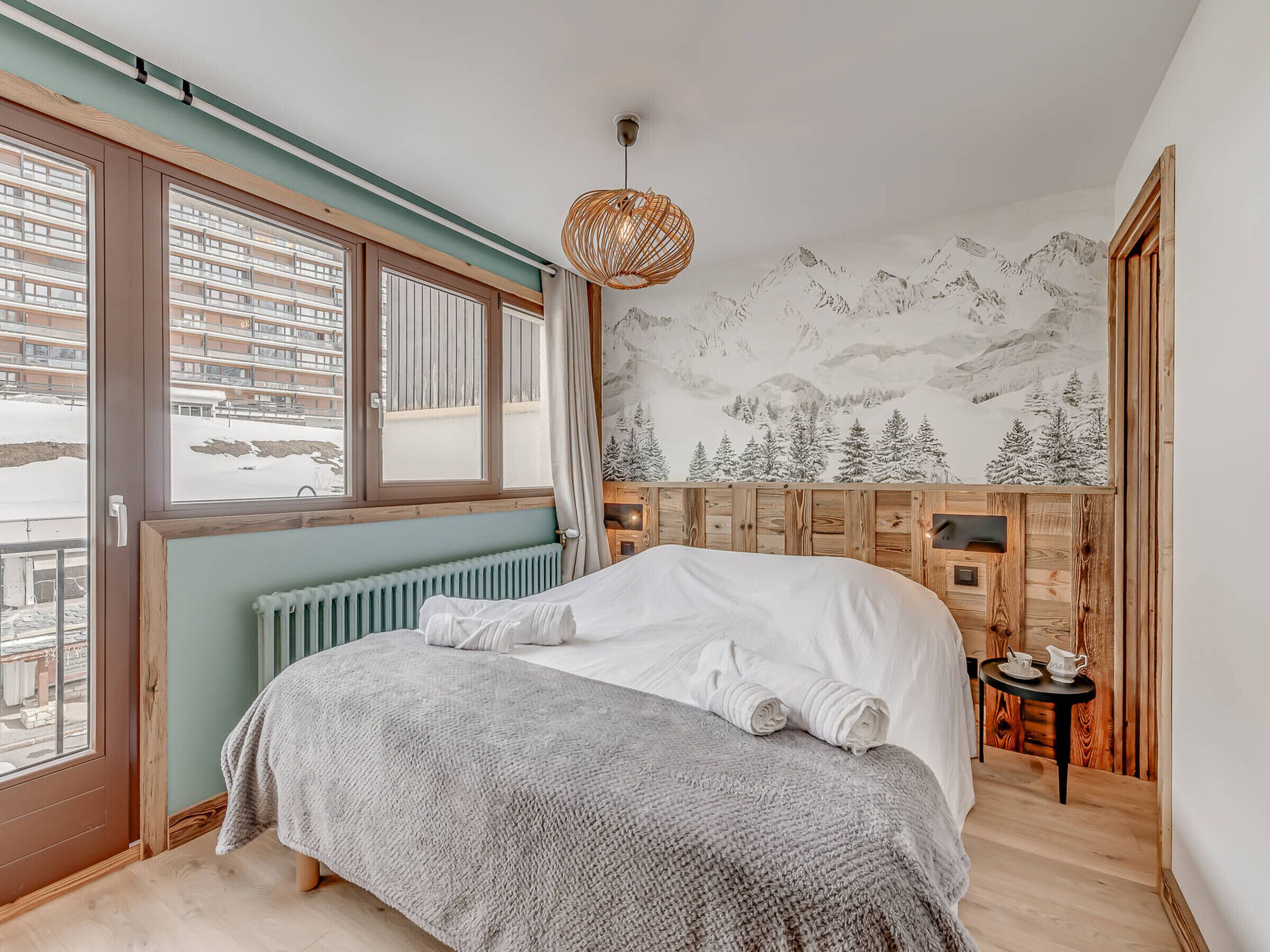 Une chambre avec un grand lit, une vue sur une montagne enneigée par la fenêtre, une tête de lit en bois et une petite table avec une théière.