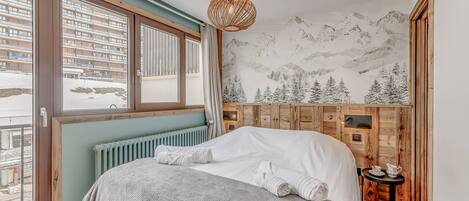 Une chambre avec un grand lit, une vue sur une montagne enneigée par la fenêtre, une tête de lit en bois et une petite table avec une théière.