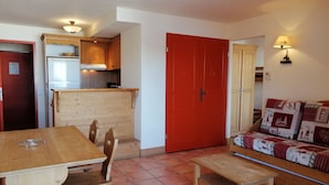 A compact living space with a wooden dining table and chairs, a red door, a kitchenette with wooden cabinets, and a sofa with patterned cushions.