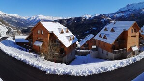 A row of wooden cabins with pitched roofs, set against a backdrop of snow-covered mountains and clear blue sky.