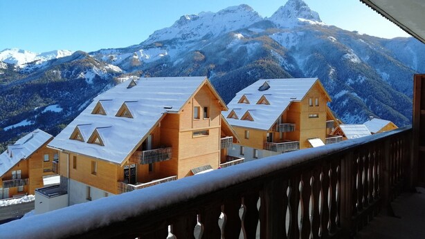 A row of wooden chalet-style buildings with snow-covered roofs, set against a backdrop of snow-capped mountains.