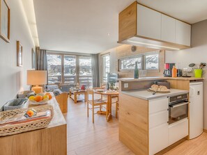 Uma cozinha moderna com armários de madeira, uma sala de jantar com mesa e cadeiras, e vista para uma paisagem montanhosa coberta de neve através de grandes janelas.