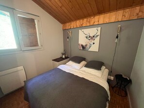 A bedroom with a bed, a nightstand, a wall-mounted lamp, a window, and a wooden ceiling.