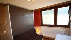 Habitació compacta amb un sofà marró fosc, una petita taula de nit amb una làmpada, una finestra amb cortines vermelles i un radiador blanc.