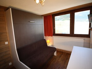A compact room with a dark brown sofa, a small bedside table with a lamp, a window with red curtains, and a white radiator. - Cozy studio in Tignes, 2 ppl, near mountains (Tignes)