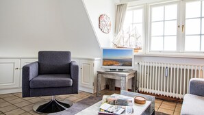 Un salón moderno con un sillón gris, un televisor de pantalla plana sobre una mesa de mármol, un modelo de barco y vistas al mar a través de amplias ventanas.