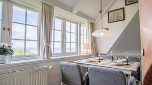 Un comedor con una mesa dispuesta para cuatro, ventanas amplias con cortinas, un radiador y cuadros en la pared.