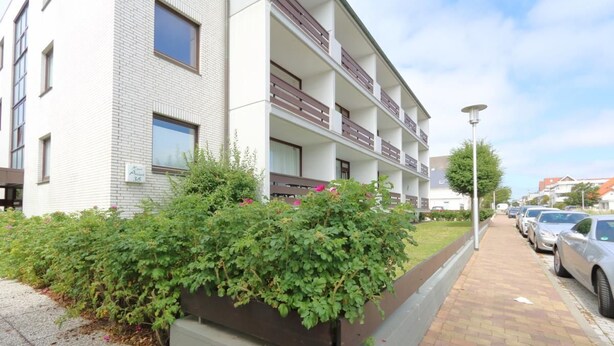 A modern apartment building with multiple balconies, a paved sidewalk, and a row of parked cars. - App. Arius 10, Germany (Sylt)