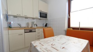 A compact kitchen with white cabinets, a microwave, a coffee maker, and a table with a coffee-themed tablecloth. - App. Arius 10, Germany (Sylt)
