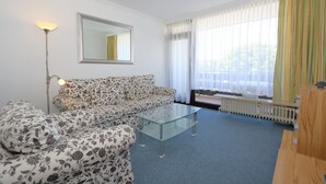 A living room with a patterned sofa, a glass coffee table, a floor lamp, and a large window with curtains. - App. Arius 10, Germany (Sylt)