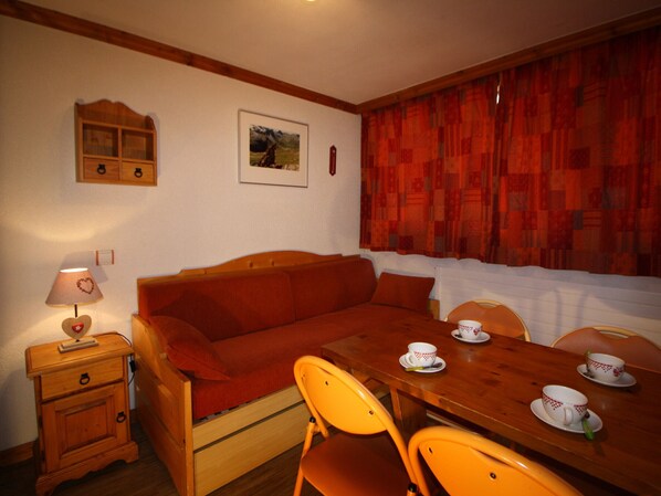 A cozy room with a wooden sofa, a small wooden table with two chairs, a nightstand with a lamp, and a window with red curtains.