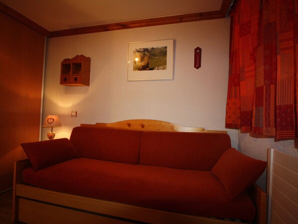 Une pièce chaleureuse avec un canapé doté d’une tête de lit en bois, un tableau représentant un paysage encadré, une horloge rouge accrochée au mur et une lampe en forme de cœur posée sur une table de chevet.