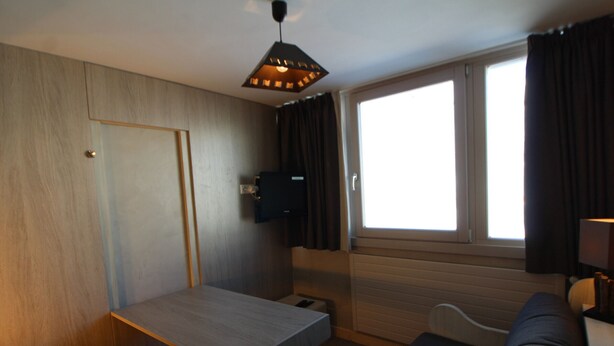 Een compacte kamer met een houten deur, een klein tafeltje, een bank, een aan de muur bevestigde televisie en een raam met gordijnen.