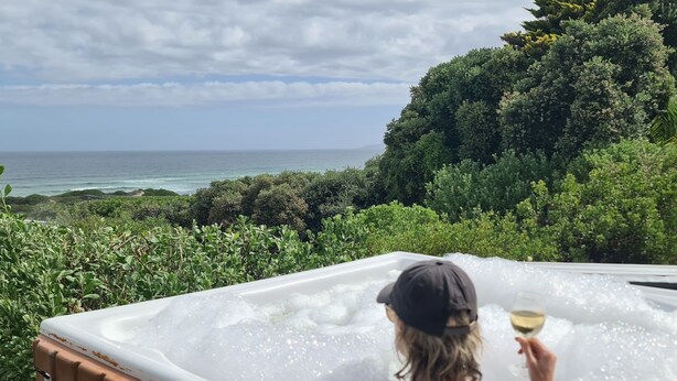 A person relaxing in a hot tub with a view of the ocean and greenery.
