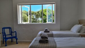 A room with two beds, a window with a view of trees, a blue chair, and a small wooden table.