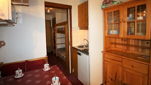 Una cocina compacta con armarios de madera, un lavamanos y una estufa. Hay un comedor con una mesa preparada para dos personas y un mantel rojo.