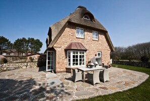 Una casa con techo inclinado, amplias ventanas y un patio de piedra con mobiliario exterior.