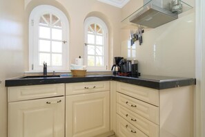 Cocina con armarios color crema, encimera negra y campana extractora de acero inoxidable. Hay dos ventanas con arcos y una cesta sobre la encimera.