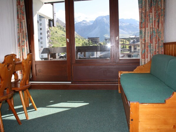 Une pièce avec un canapé et des fauteuils en bois, et un balcon offrant une vue sur les montagnes et les bâtiments.