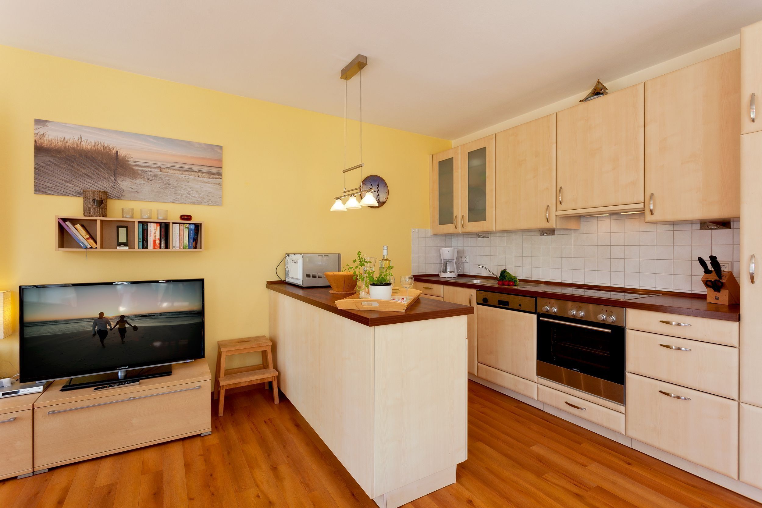 A modern kitchen with wooden cabinets, a central island, a built-in oven, and a flat-screen TV.