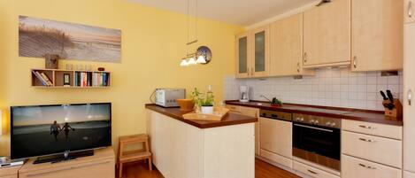 A modern kitchen with wooden cabinets, a central island, a built-in oven, and a flat-screen TV.