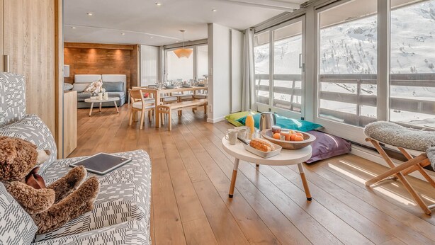 A modern living room with a wooden floor, a sofa, a dining table with chairs, and a large window with a snowy view. - Modern Apartment 3* Tignes - 80m² - 8 people (Tignes)