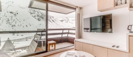 A modern living room with a large window overlooking a snowy mountain landscape, a flat-screen TV mounted on the wall, and a small wooden table with magazines.