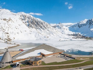Paisagem nevada de montanhas com um lago, uma estrutura de madeira com telhado cônico e um edifício com detalhes em laranja.