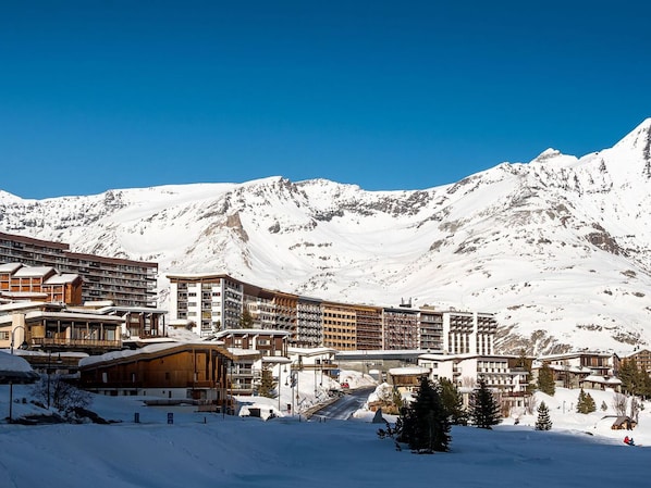 A snow-covered mountain resort with multi-story buildings, ski slopes, and a clear blue sky.