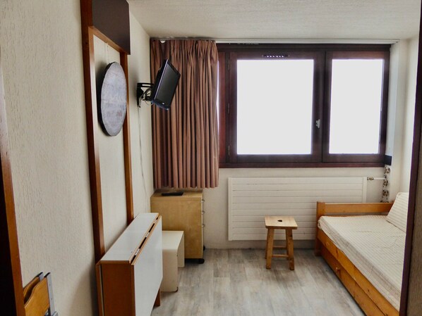 A compact room with a bed, a small table, a desk, a chair, a television, and a window with curtains.