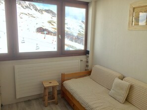 Un dormitorio con una cama de madera, un taburete pequeño de madera, un radiador y una ventana que ofrece vistas a una montaña nevada y un telesilla.