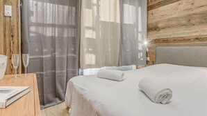 A neatly made bed with white linens, a wooden nightstand with a glass, and a window with sheer curtains.