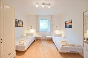 Quarto com duas camas de solteiro, uma pequena mesa, janela com cortinas e obras de arte nas paredes.