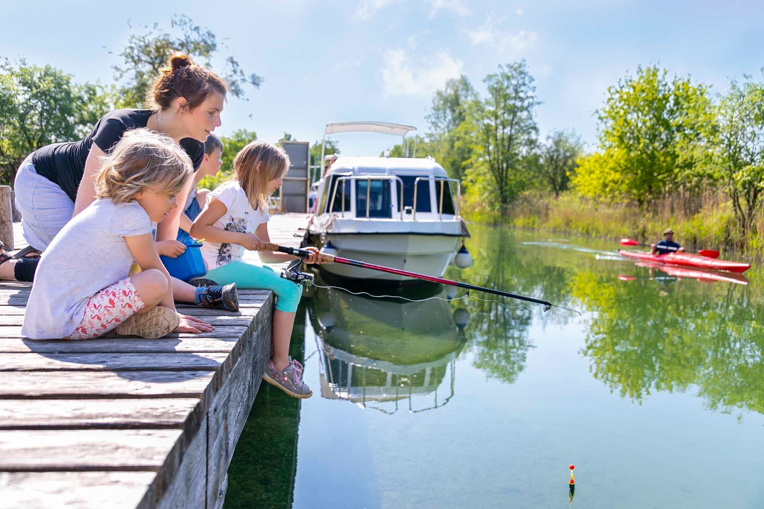 A wooden dock with people fishing by a calm river, a boat docked nearby, and lush greenery in the background.