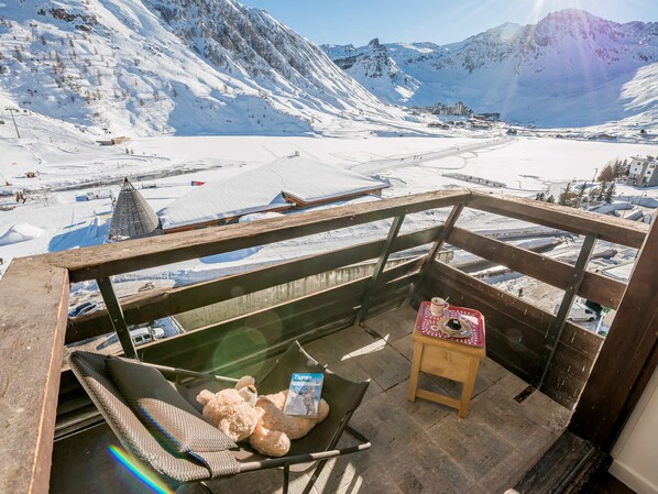 Un balcon donnant sur un paysage montagneux enneigé, une chaise, une petite table avec une tasse et un livre, et une balustrade en bois.