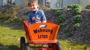 Een kind in een rode wagentje, met daarop 'Wohnung LITUS' geschreven.